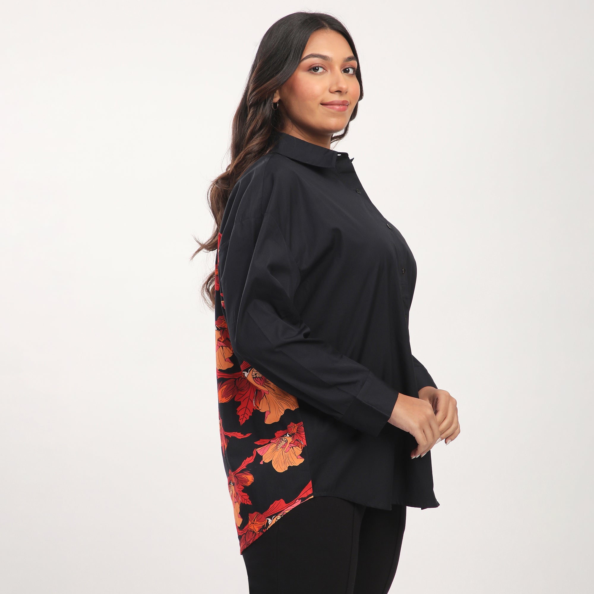A woman modeling a plus size black cotton floral print color block poplin shirt with long sleeves, shown from the back, with a red handbag and a rose flower accessory.