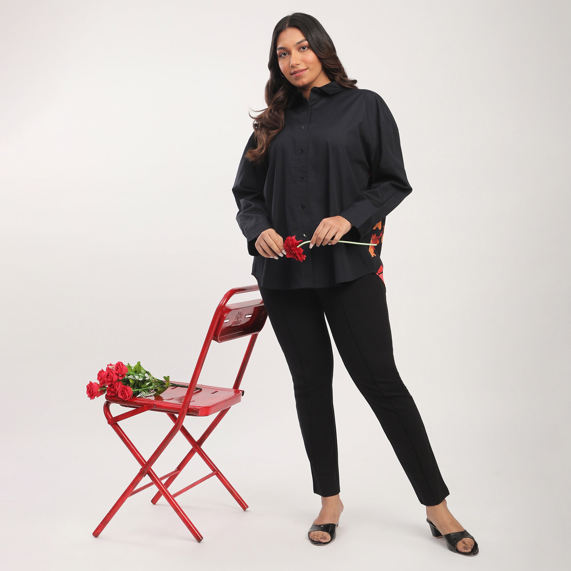 Woman in black outfit holding a red rose next to a red chair on a white background