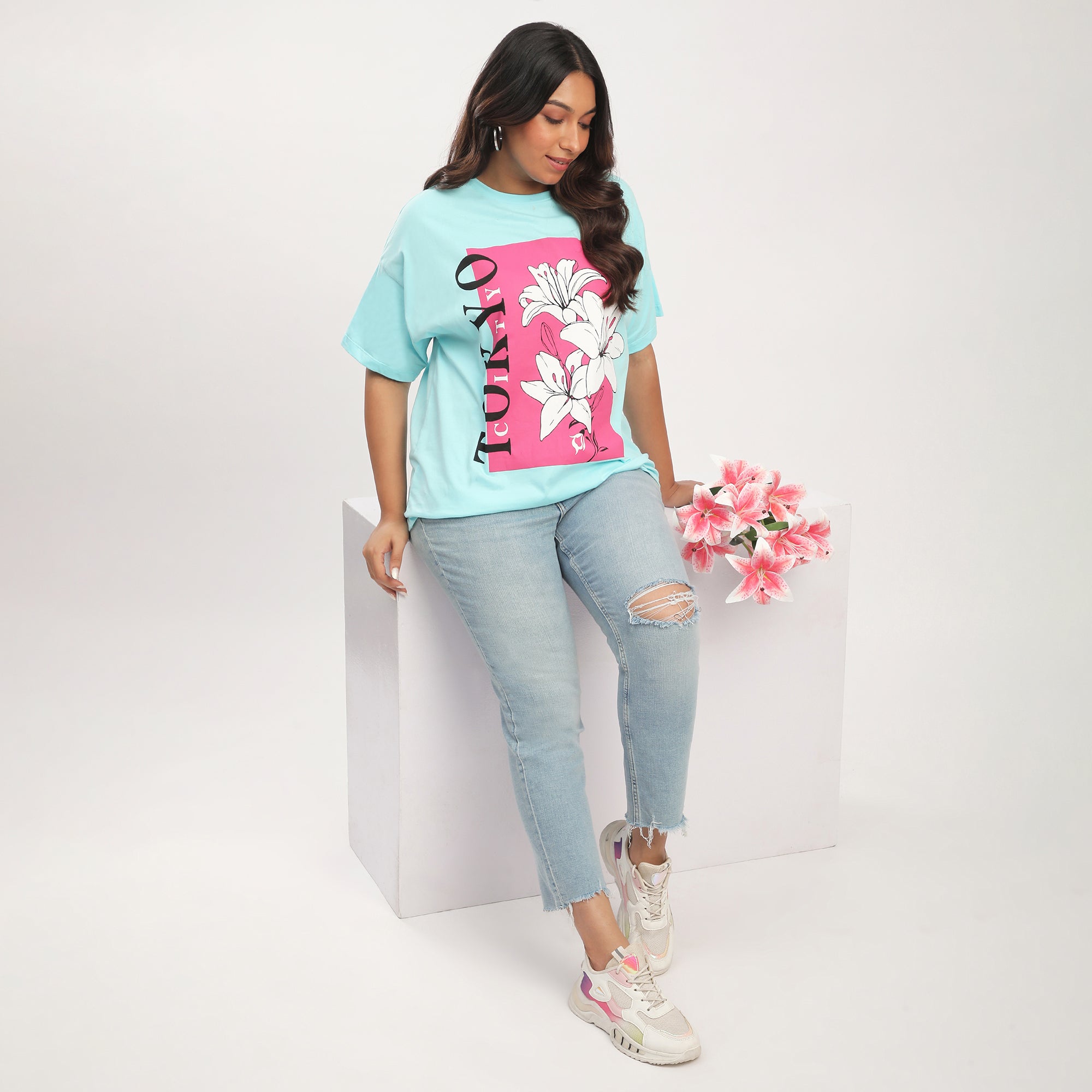Woman in light blue t-shirt with floral design and ripped jeans standing next to a white box with pink flowers.