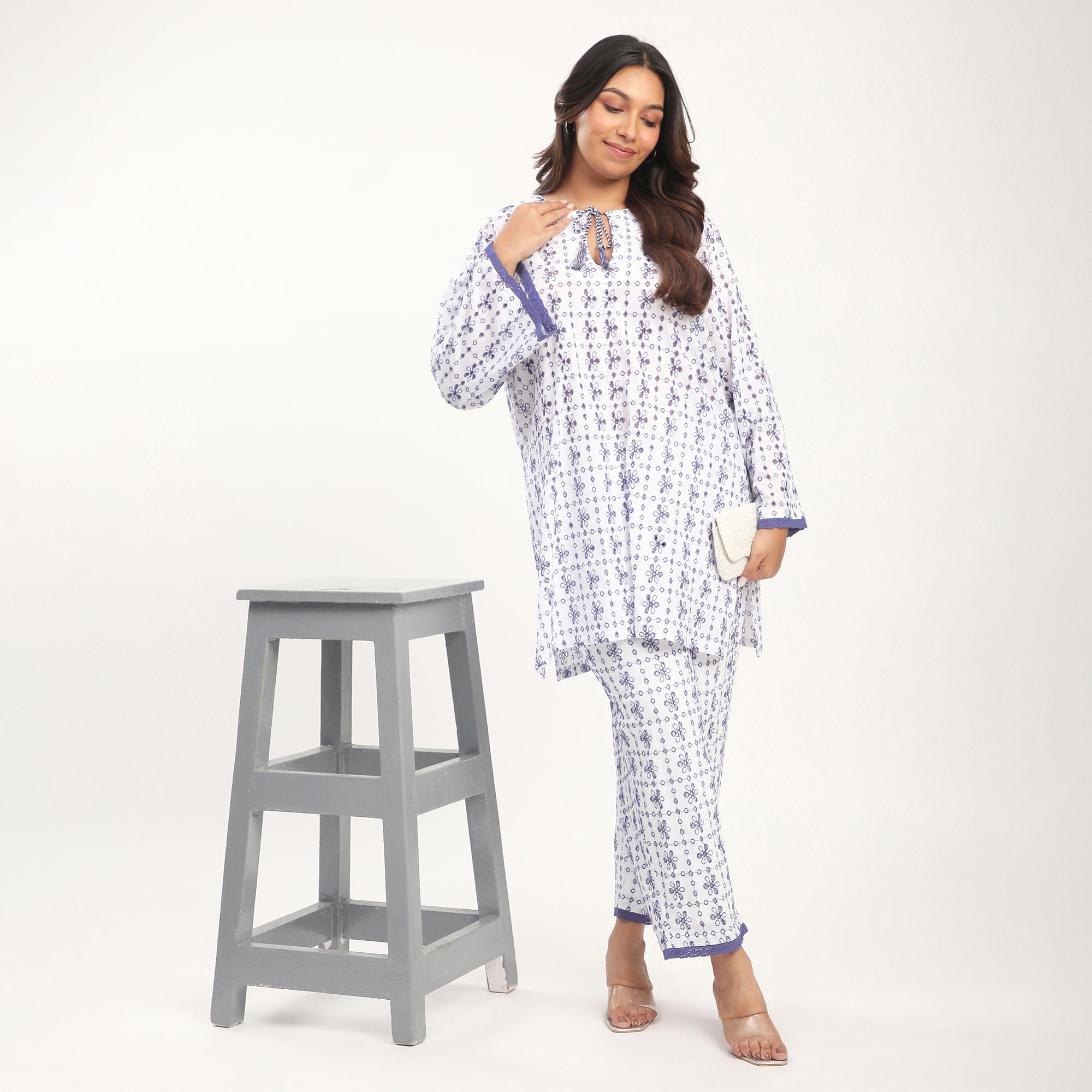 Woman wearing a patterned Cotton Schiffli Cutwork Lace Detail Co-ord Set standing next to a gray stool on a white background.
