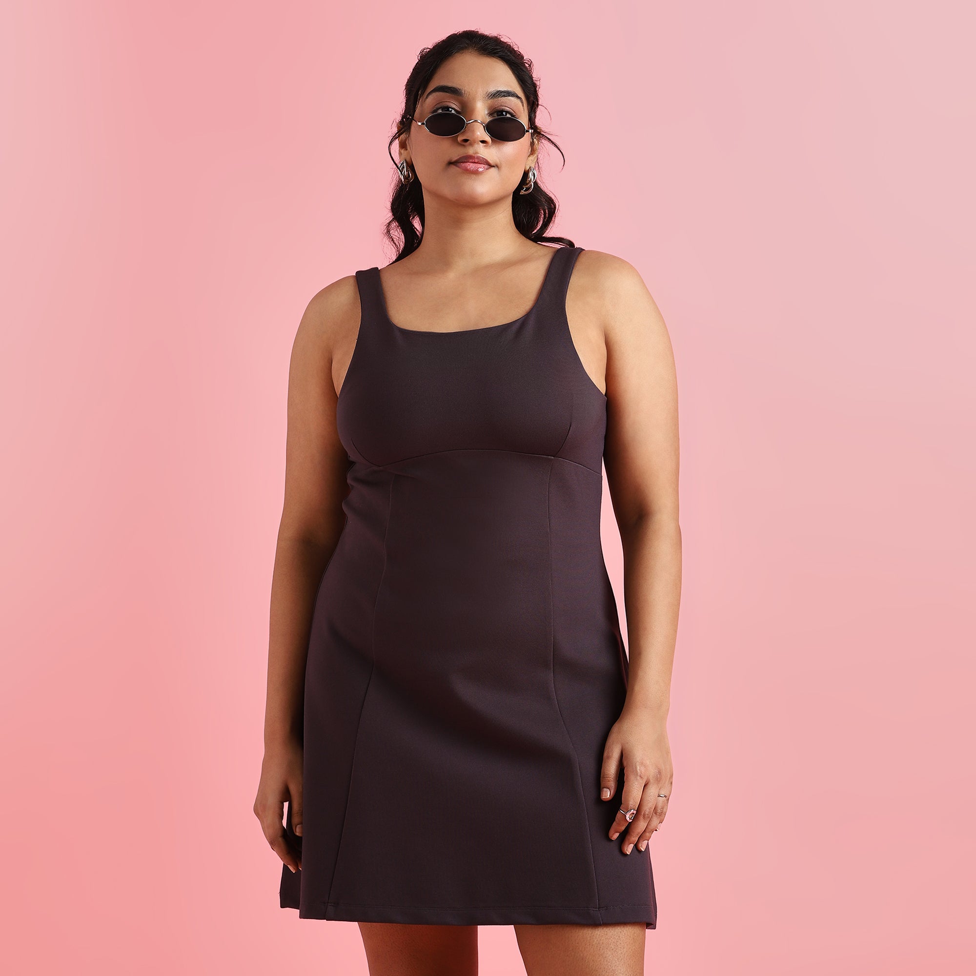 A plus-size woman standing against a pink background, wearing a charcoal gray square neck 4-way stretch skater dress with black ankle boots.