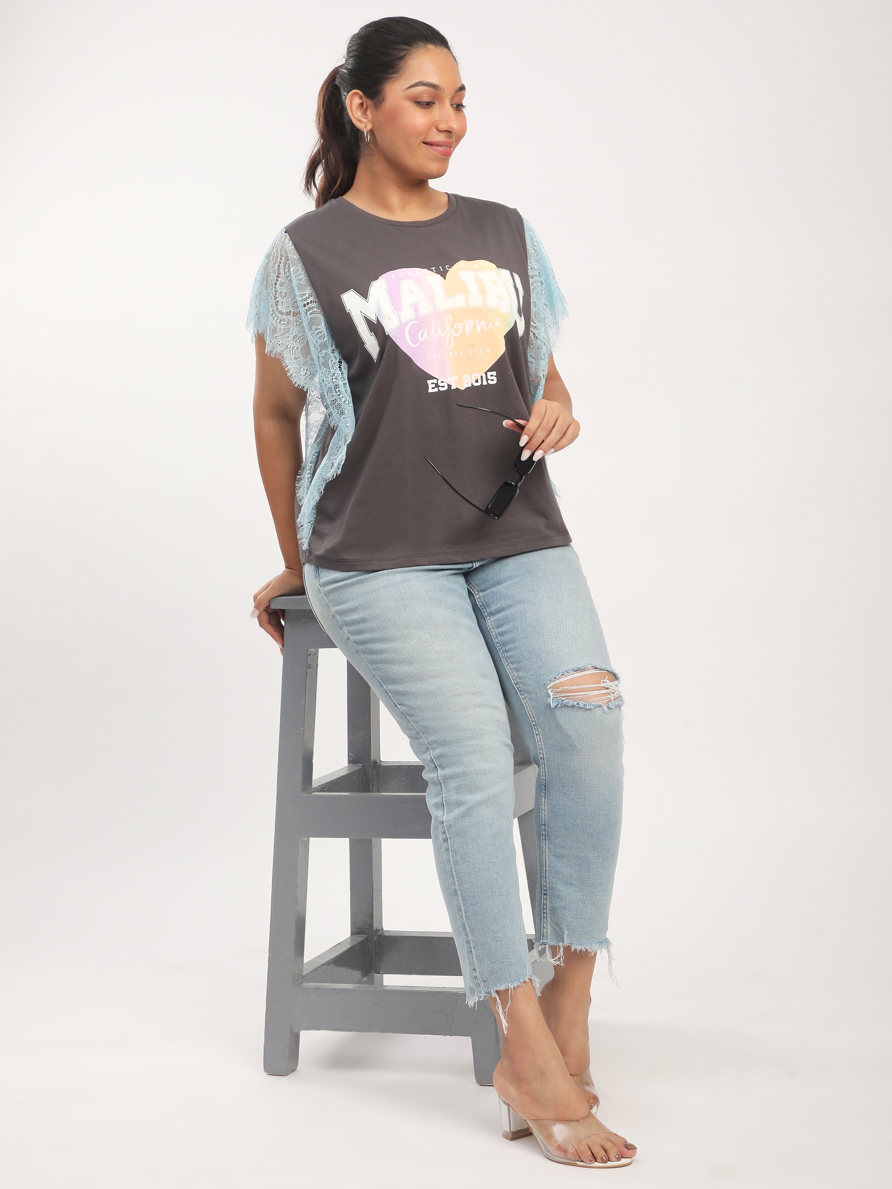 Woman in graphic t-shirt and jeans sitting on a stool against a plain background