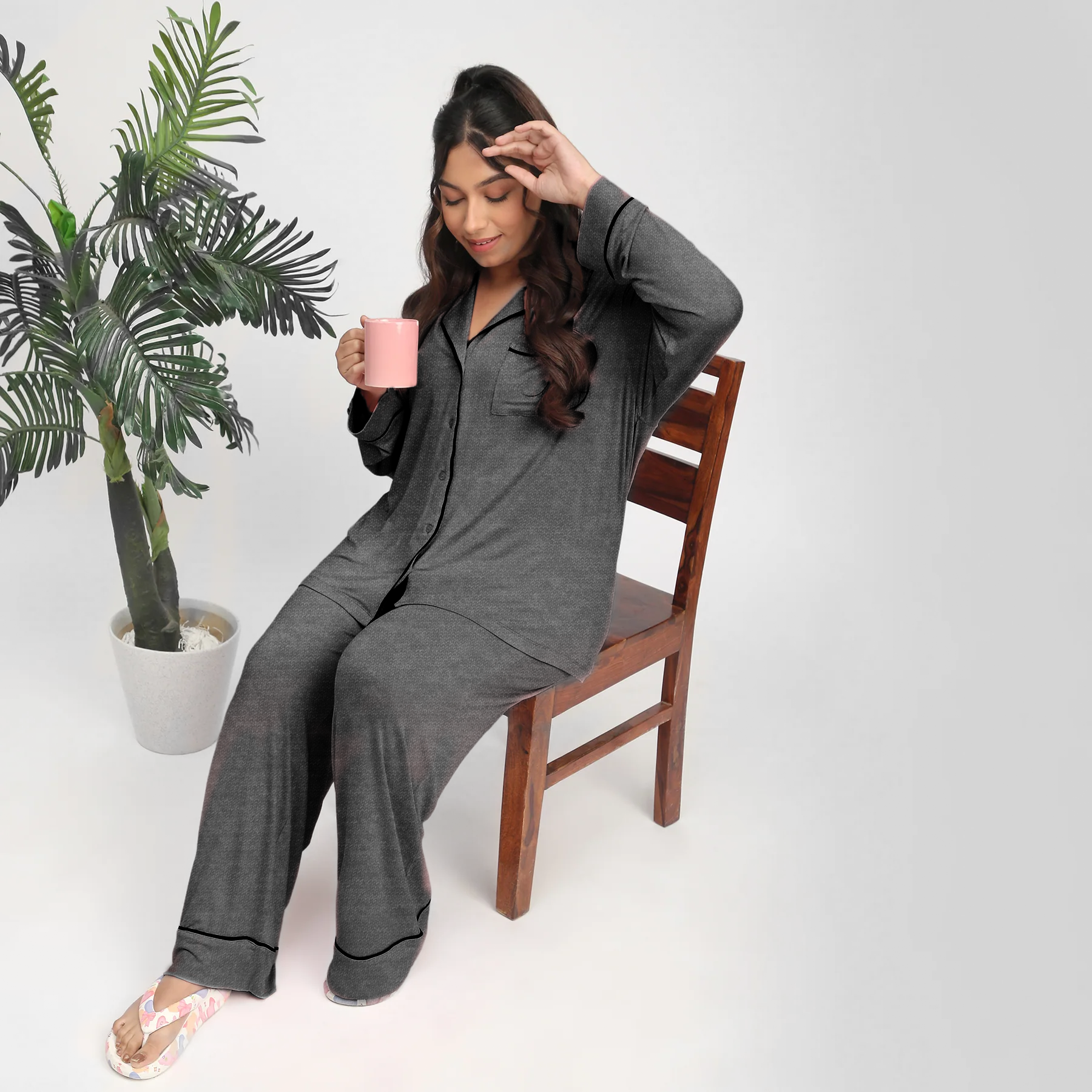Woman in gray pajamas sitting on a chair holding a pink mug with a plant in the background.
